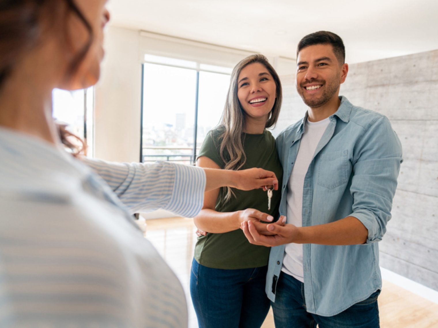 A happy couple is receiving keys from a real estate agent, as they stand in a bright, modern apartment.