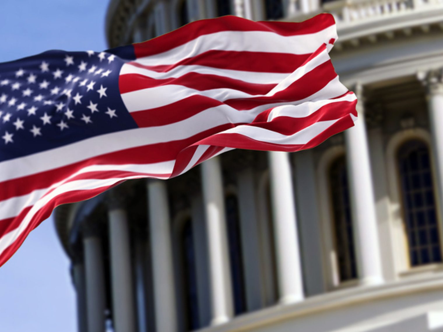 The United States flag flies prominently in front of a classical government building with iconic columns, against a clear blue sky.