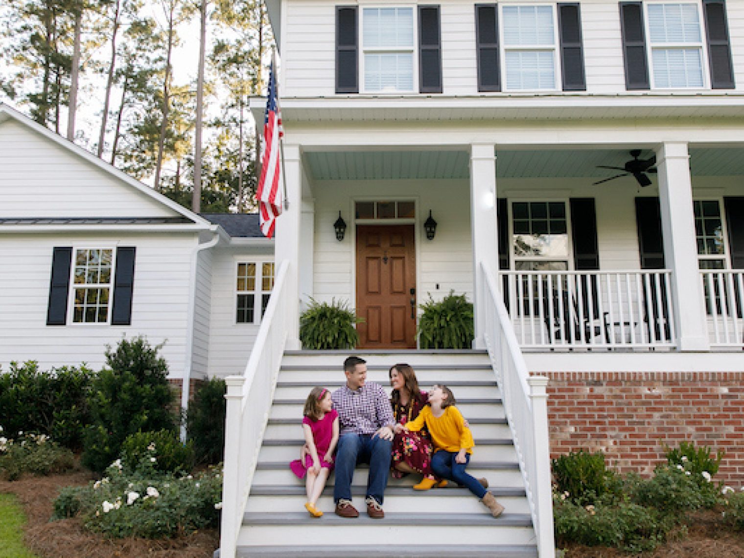 Family sitting outside on the steps of a new construction white siding farmhouse in the suburbs